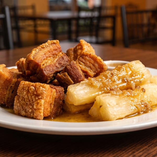Crispy pork belly chunks and yuca with a savory sauce on a white plate on a wooden table.