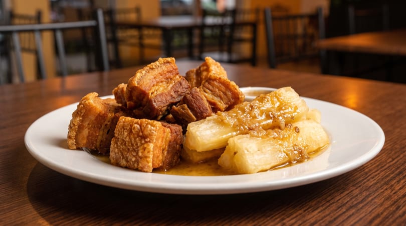 Crispy pork belly chunks and yuca with a savory sauce on a white plate on a wooden table.